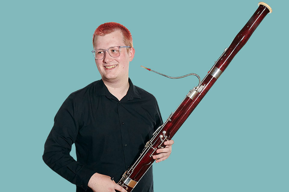 A male student wearing a black shirt, smiling, holding a bassoon.